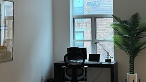 Un espacio de oficina moderno con un escritorio, una silla, una computadora y un amplio ventanal con vista a un edificio de ladrillo.