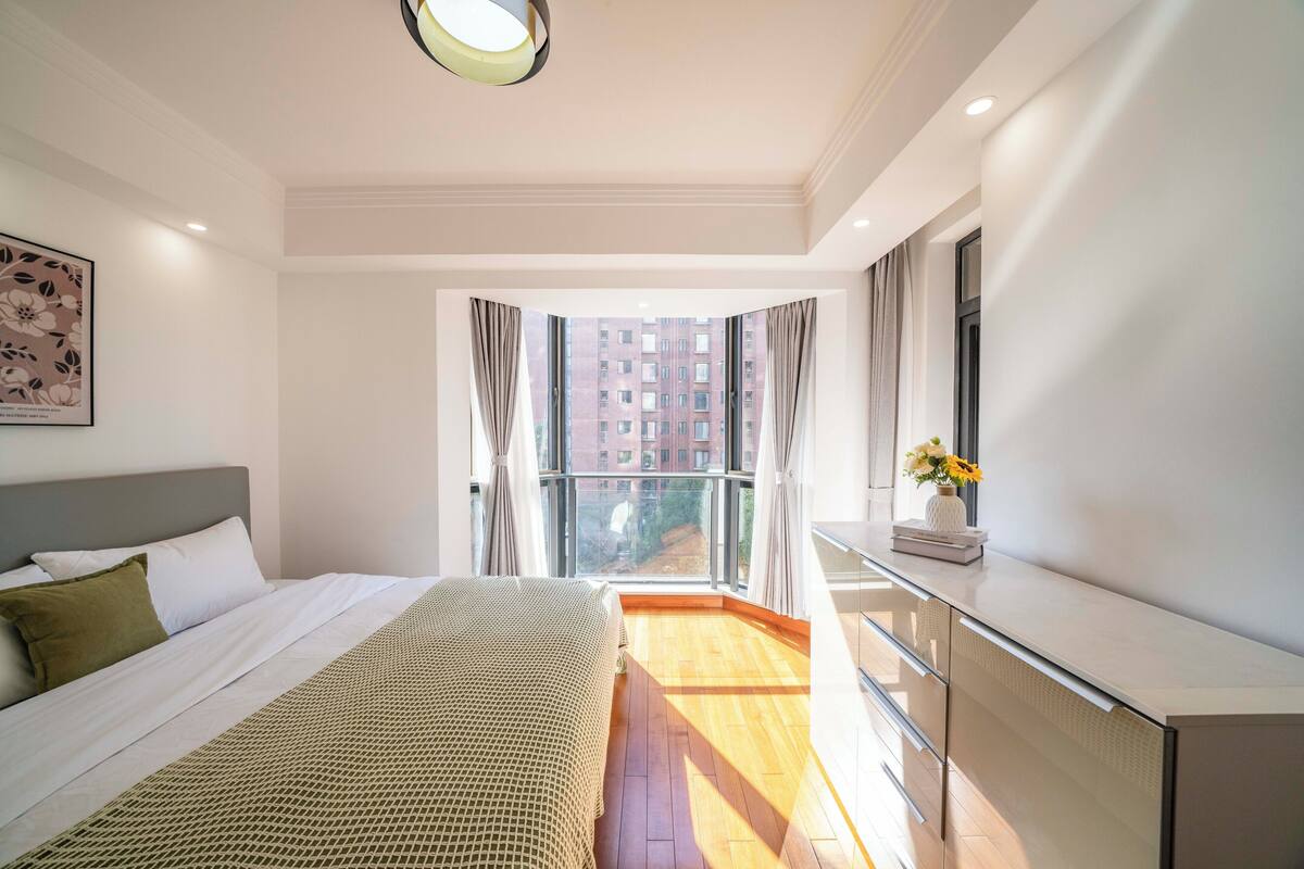 A bedroom with a bed, a dresser, a window with curtains, and a view of buildings.