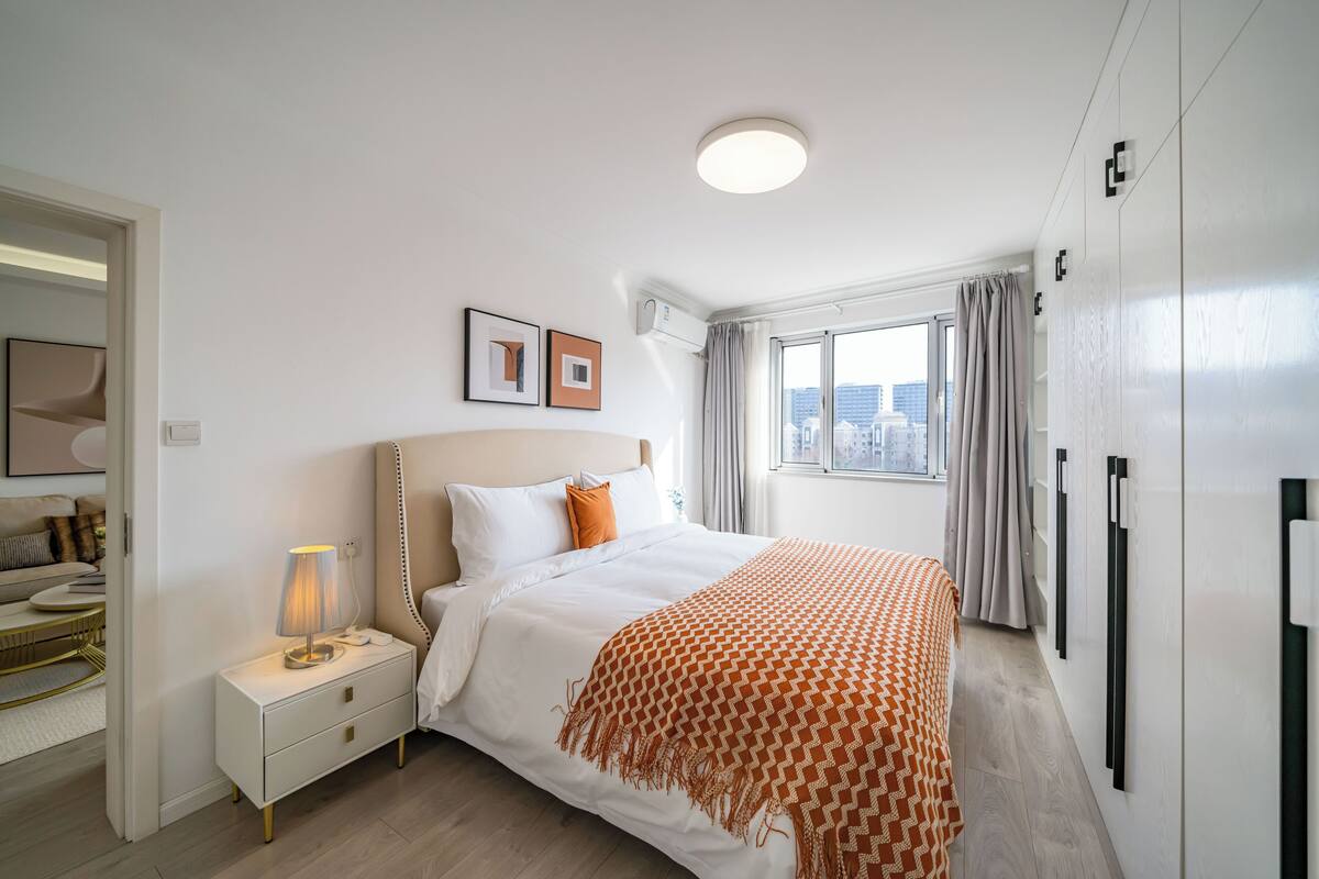 Een moderne slaapkamer met een groot bed, een nachtkastje met een lamp, een raam met gordijnen en uitzicht op de stad.