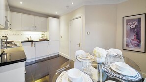 A modern kitchen with white cabinets, a black countertop, and a dining area with a glass table set for four. - Central Mayfair Luxury Apartments (London)