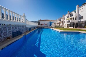 A swimming pool with a white tiled edge, a white fence with decorative elements, and a building with a tiled roof and balconies. - Summer Azul 3 Casa Lena by Casasol (Nerja)