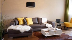Una sala de estar moderna con un sofá marrón oscuro, cojines amarillos, una manta suave de color blanco, una mesa de centro de vidrio con velas y una lámpara de pie.