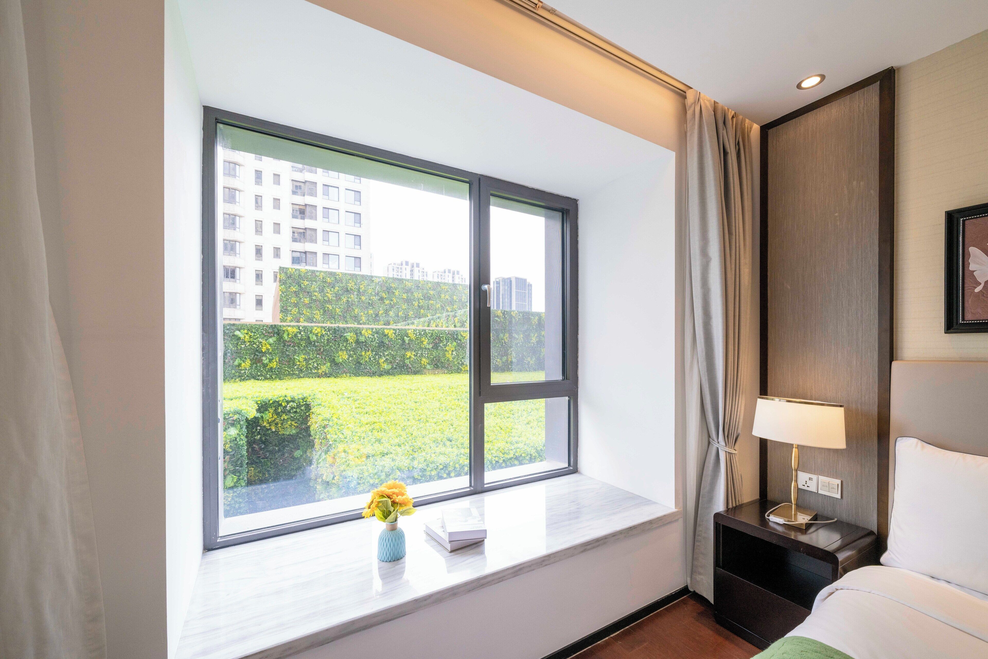 A hotel room with a large window, a bedside table with a lamp, a bed, and a view of a green landscape and buildings outside.