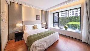 A hotel room with a large bed, two bedside tables with lamps, a window with a view of a building and greenery, and a wall-mounted air conditioner. - Tyms Gubei Apt 2BR Apt Near Takashimaya (Shanghai)