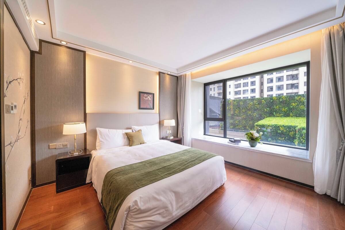 A hotel room with a large bed, two bedside tables with lamps, a window with a view of a building and greenery, and a wall-mounted air conditioner.
