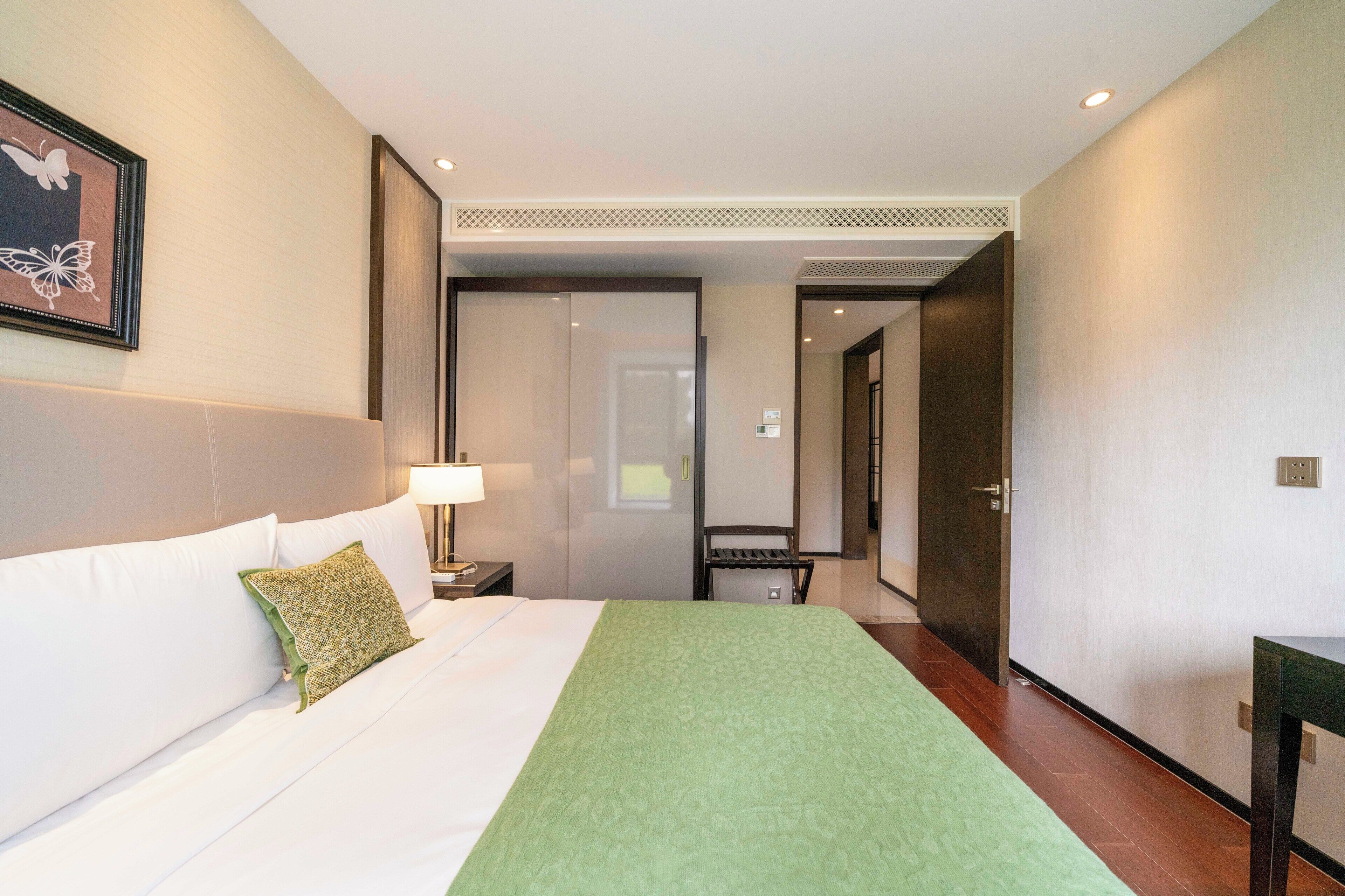 Ein Hotelzimmer mit einem Bett, einem grünen Überwurf, einem Nachttisch mit Lampe und einem gerahmten Schmetterlingsbild an der Wand.