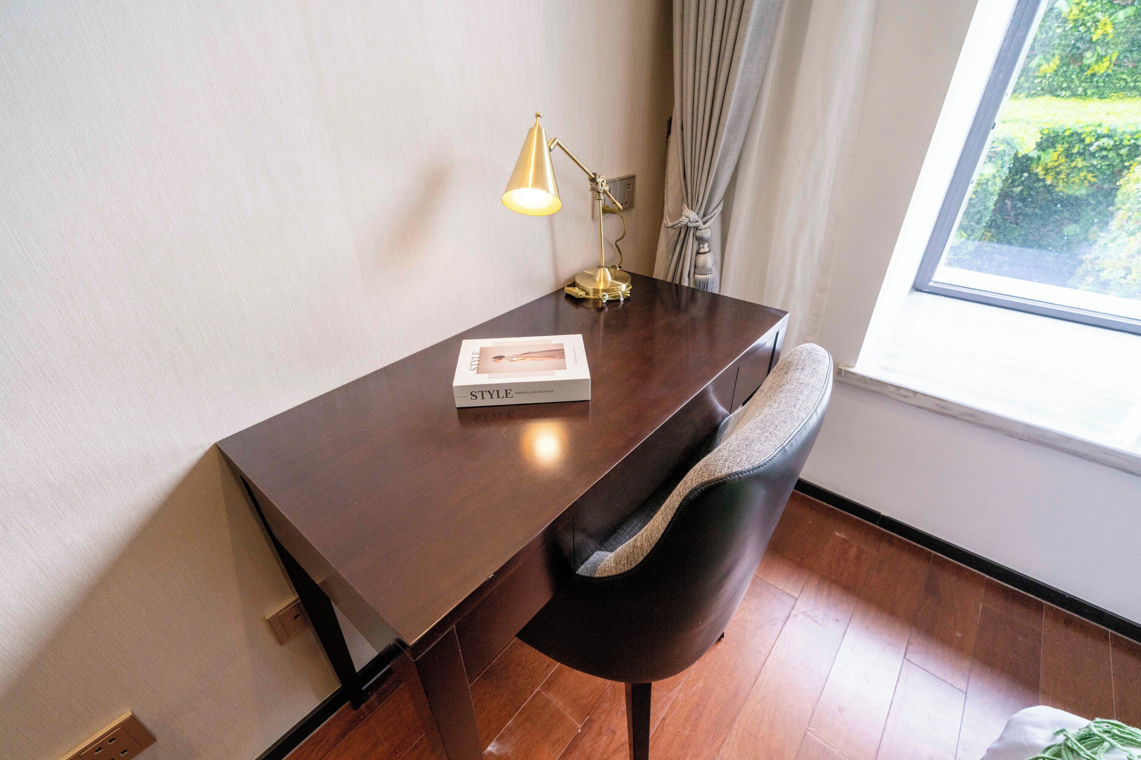 Un bureau en bois, une lampe, une chaise et un livre intitulé « STYLE » se trouvent dans une pièce dotée d’une fenêtre et de rideaux.