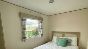 Ein Schlafzimmer mit einem Einzelbett, einem Nachttisch, einem Fenster mit Vorhängen und Blick auf einen grünen Zaun und Bäume.