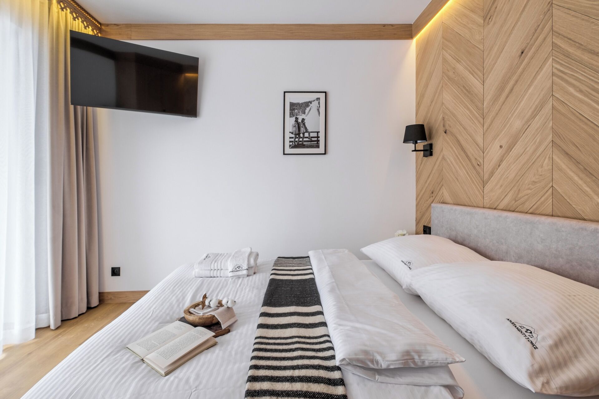 A bedroom with a bed, a flat-screen TV mounted on the wall, a framed picture, and a wooden headboard.