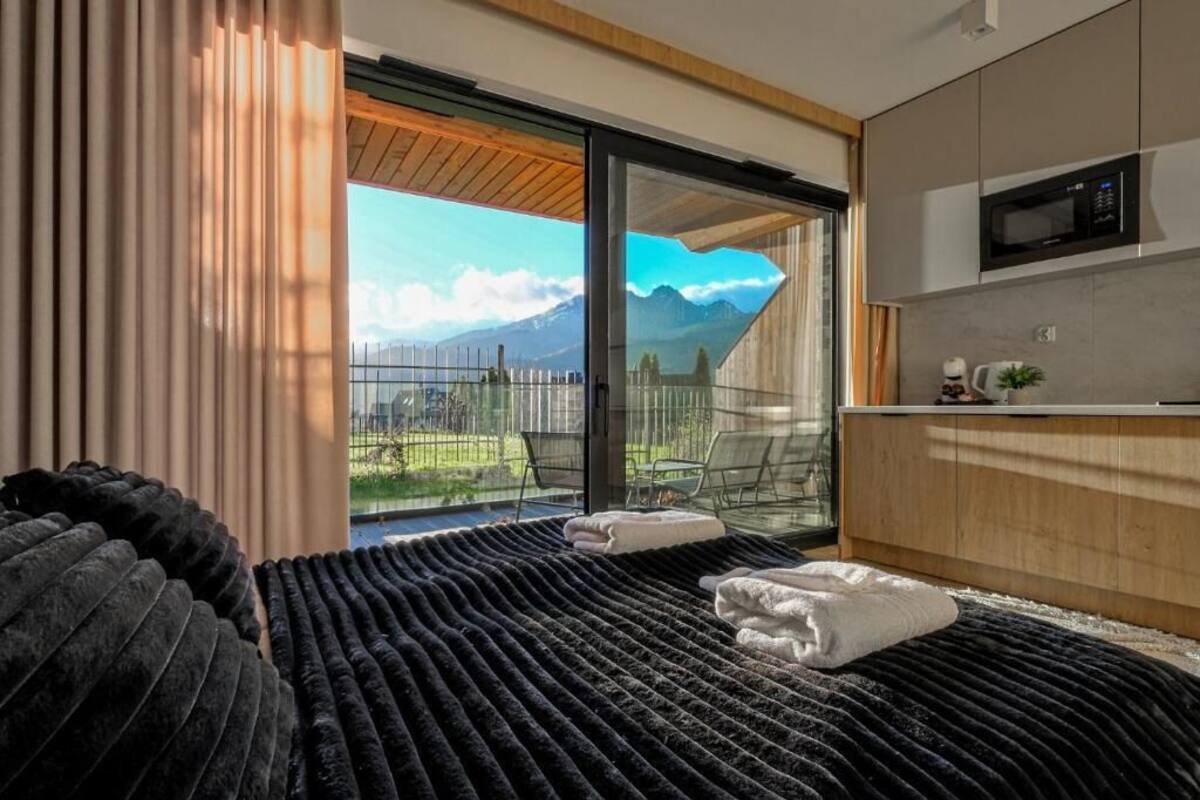 A modern hotel room with a large bed, a view of mountains, a balcony with a chair, and a kitchenette.