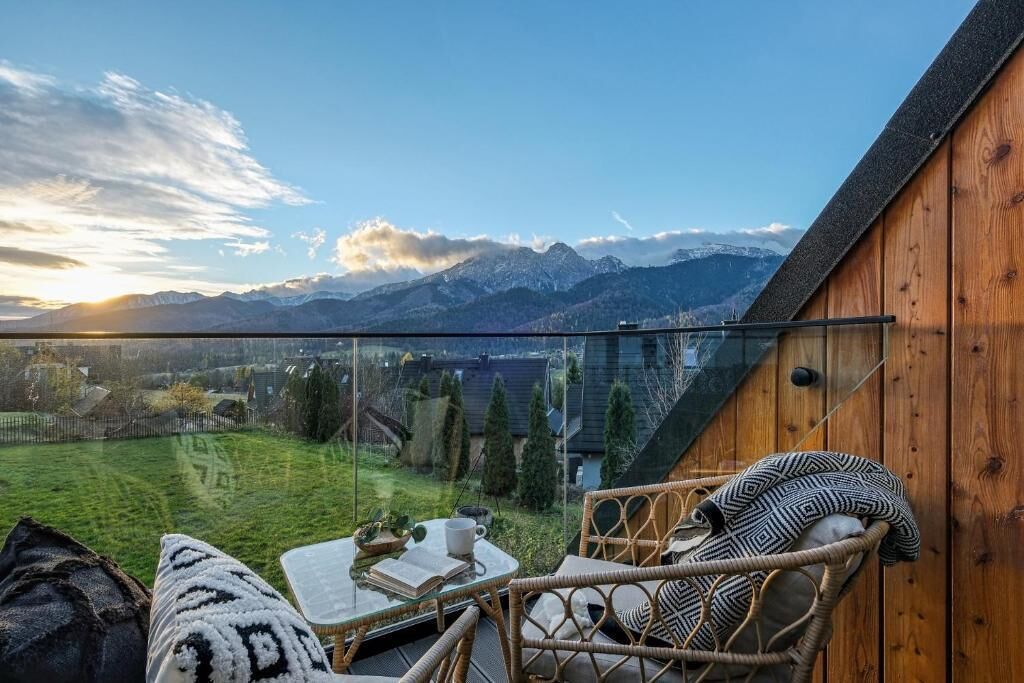 Ein Balkon mit Bergblick, ein Korbstuhl, ein Tisch mit einem Buch und einer Tasse sowie eine Holzwand.