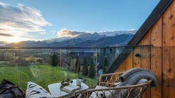 Ein Balkon mit Bergblick, ein Korbstuhl, ein Tisch mit einem Buch und einer Tasse sowie eine Holzwand.
