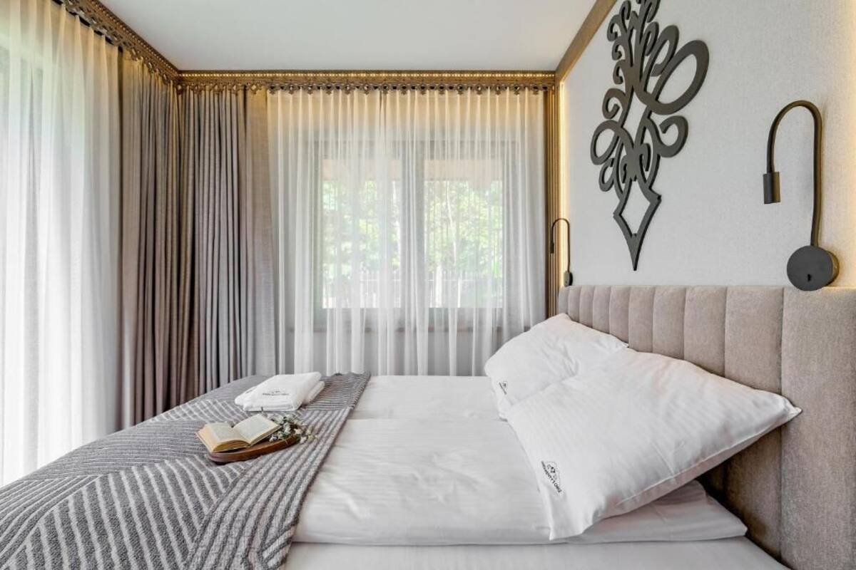 A hotel room with a large bed, a bedside table with a book and a glass, sheer curtains, and a decorative wall piece.