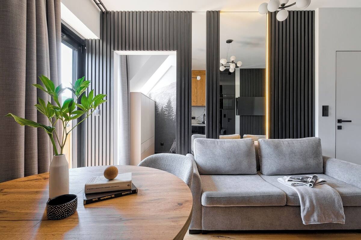 A modern living room with a grey sofa, a round wooden table with a plant and books, and a view of the kitchen and outside.