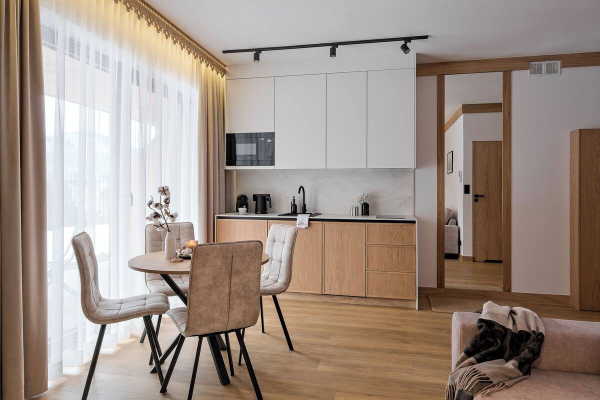 A modern kitchen with white cabinets, a black microwave, and a coffee machine. There is a dining area with two chairs and a table, and a living room with a sofa and a blanket.