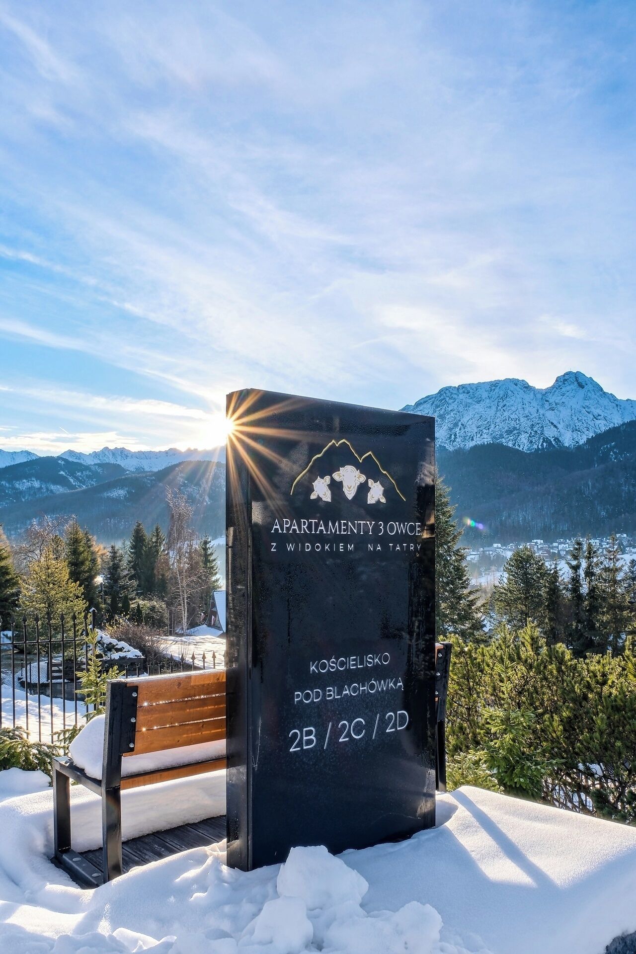 A sign for 'Apartamenty 3 Owcze' with a view of snow-covered mountains and a bench in the foreground.
