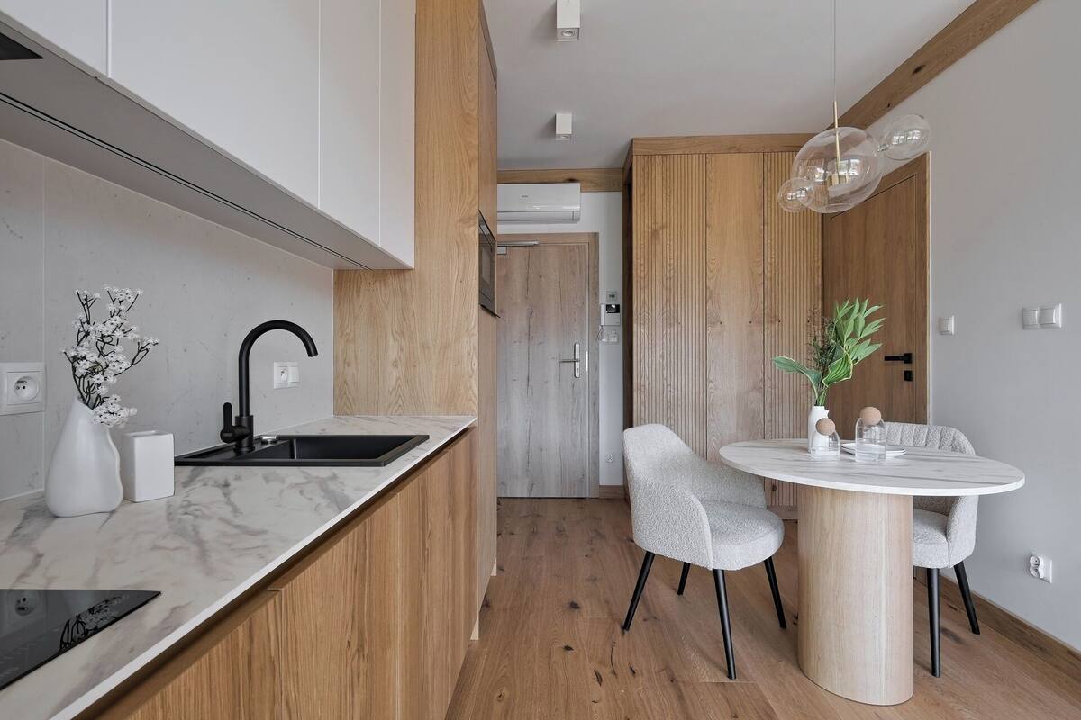 A modern kitchen with a marble countertop, black faucet, and wooden cabinetry. There is a dining area with a round table and two chairs.