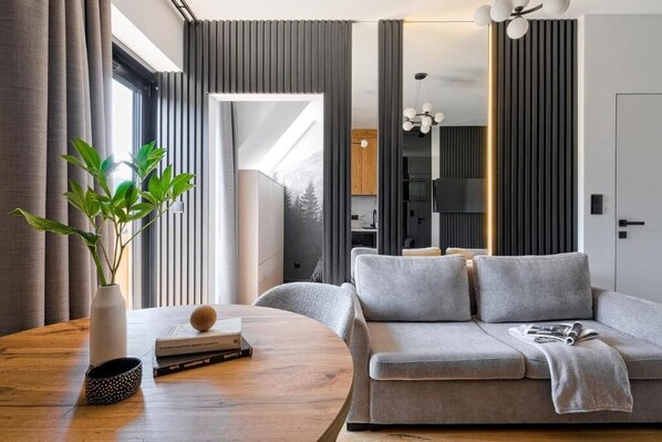 A modern living room with a grey sofa, a round wooden table with a vase and books, and a view into the kitchen area.