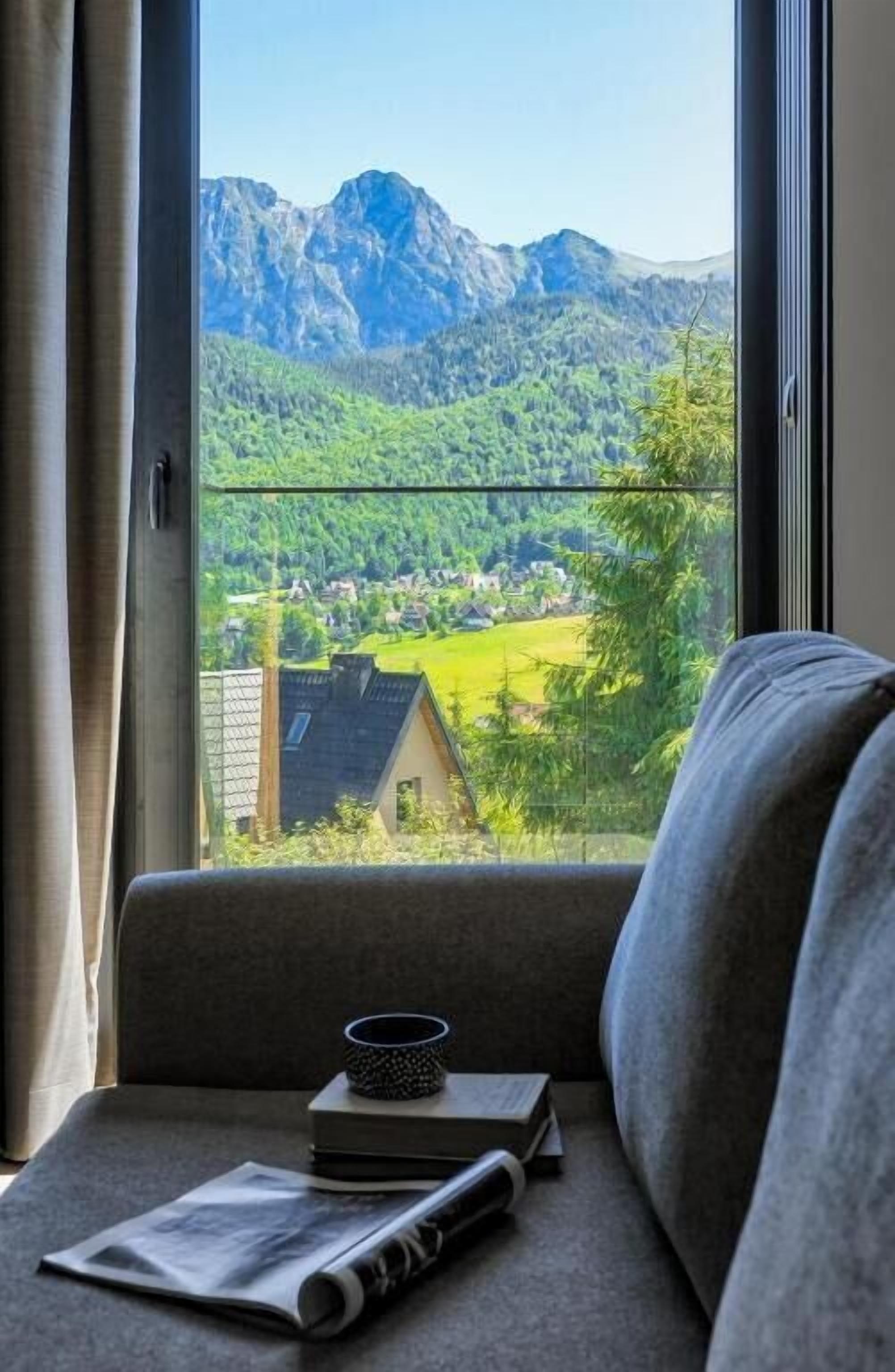 A view from a window with a mountain landscape, a sofa with a book and a cup, and a newspaper on the windowsill.