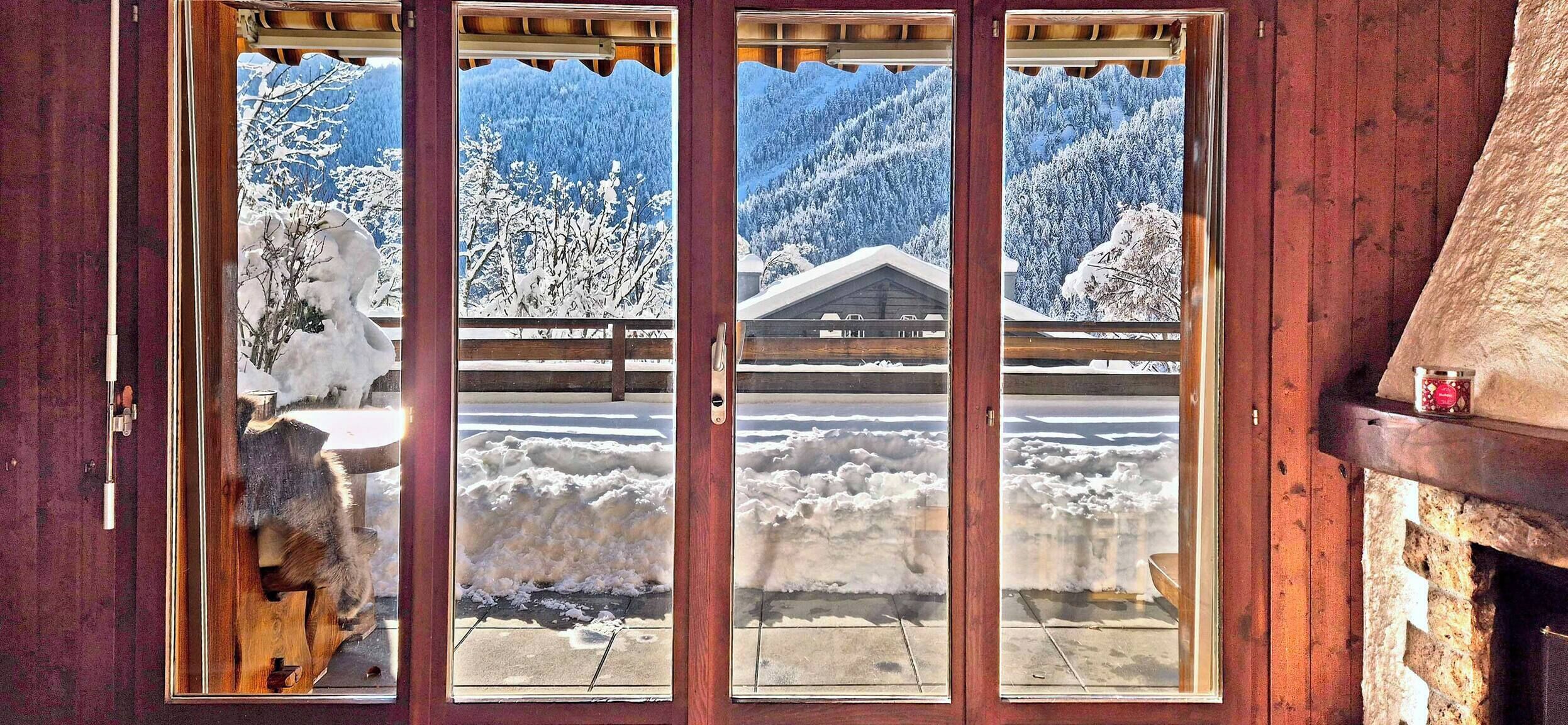 Uma porta de madeira com três painéis de vidro. O painel da esquerda mostra uma paisagem nevada com árvores e um edifício. O painel central exibe uma cadeia montanhosa coberta de neve. O painel direito apresenta uma paisagem nevada com árvores e um edifício.