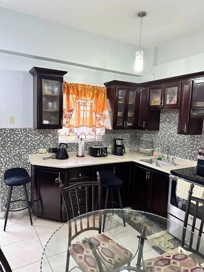 A kitchen with dark wooden cabinets, a tiled backsplash, a glass island with stools, and a window with orange curtains. - The Oasis –Kingston 5 Luxury Loft, 24hrs security (Kingston)