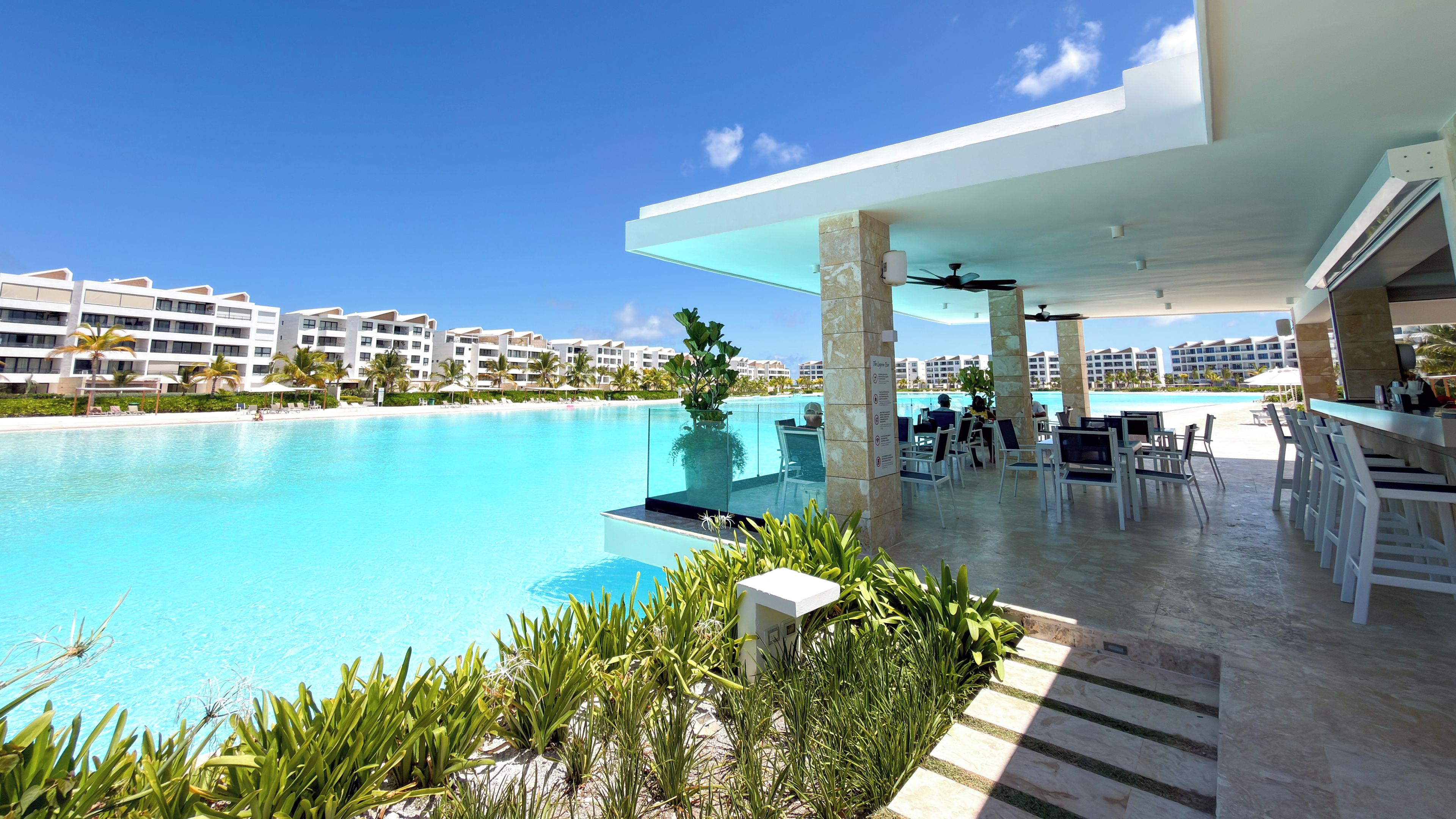 Ein Pool in einem Resort mit Bar, Liegestühlen und Blick auf den Ozean sowie weitere Gebäude des Resorts.