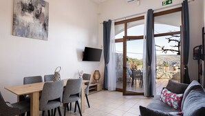 Een moderne kamer met een eettafel, grijze stoelen, een bank met bloemenkussens, een flatscreen-tv en een grote schuifdeur naar een buitenruimte met een stenen muur en een zichtbare tak.