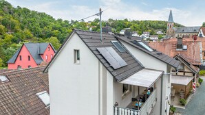 A residential building with a balcony, solar panels, and a view of a church and forested hill.