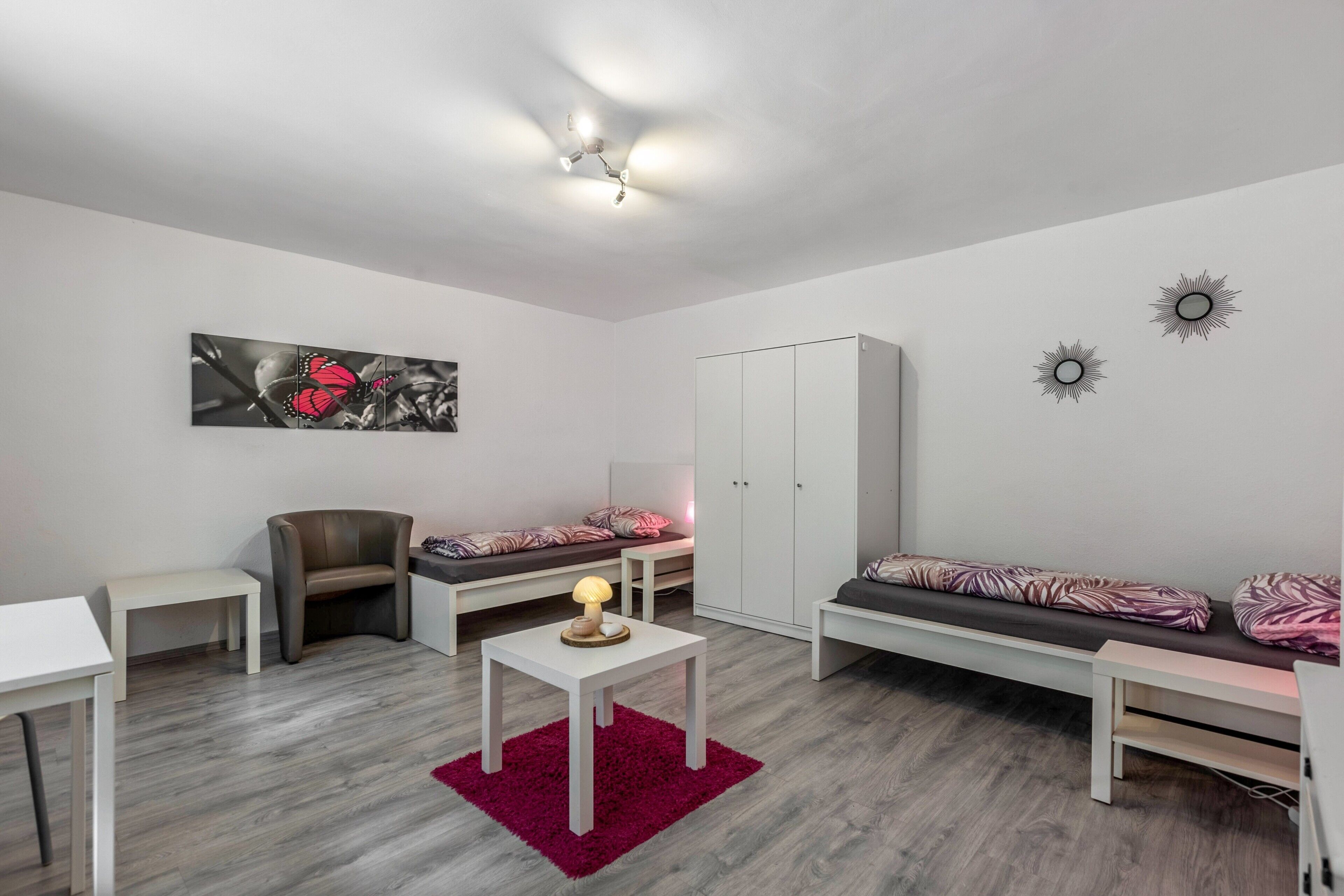 Ein modernes Wohnzimmer mit einer Couch, einem Sessel, einem kleinen Tisch mit Lampe, einem Bett mit gemusterten Betttextilien und einem Kleiderschrank.