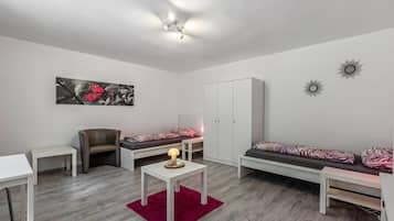 Ein modernes Wohnzimmer mit einer Couch, einem Sessel, einem kleinen Tisch mit Lampe, einem Bett mit gemusterten Betttextilien und einem Kleiderschrank.