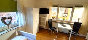 A room with a bed, a desk, a chair, a TV, and a window with a view of the outside.