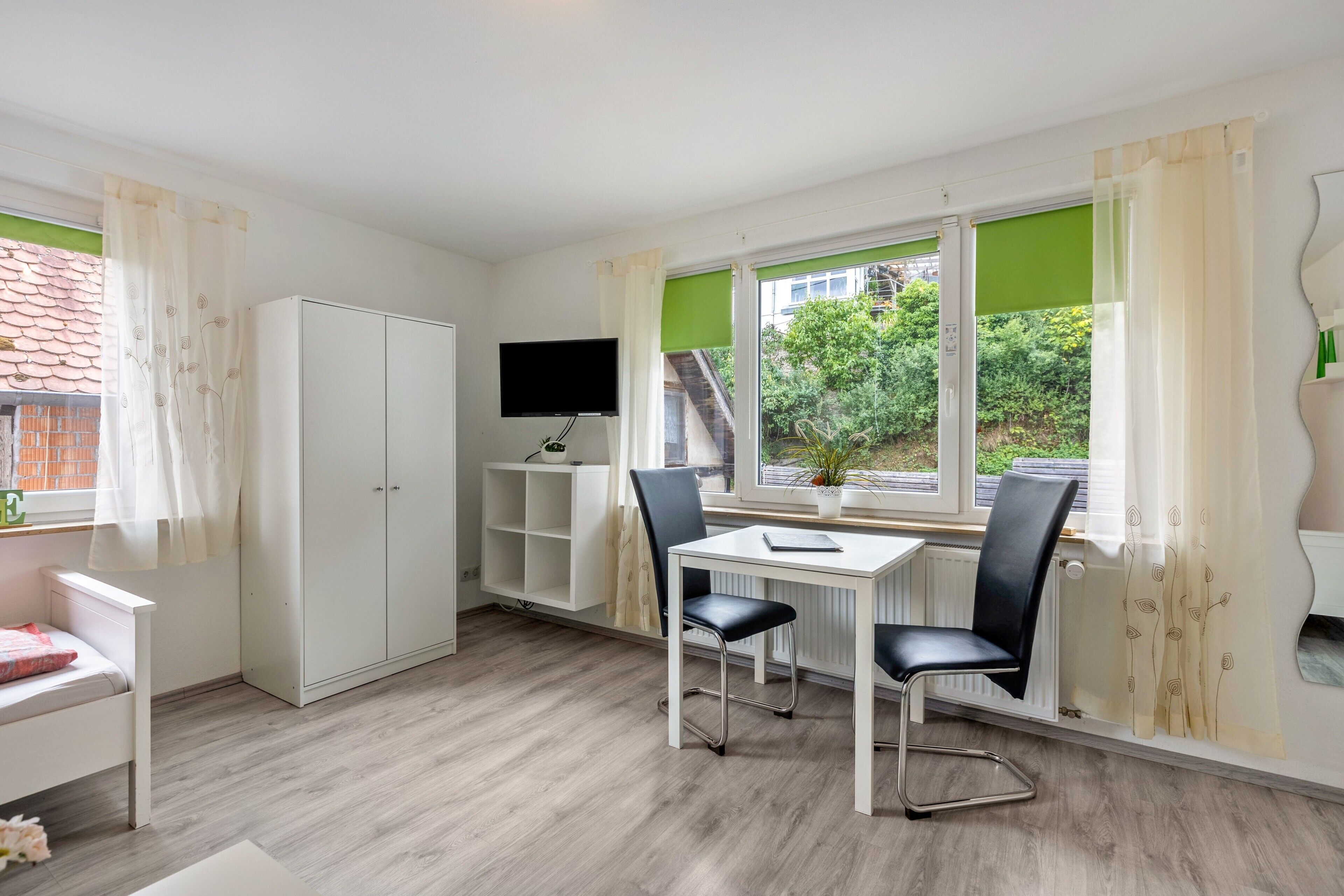 Ein Zimmer mit einem weißen Kleiderschrank, einem Schreibtisch mit zwei Stühlen, einem Fernseher und einem Fenster mit Blick auf Grünflächen.