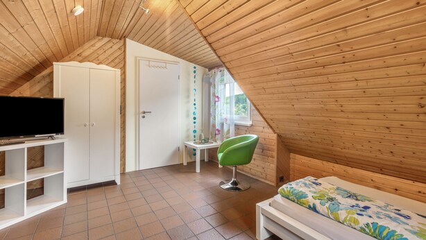 A small, sloped-roofed room with a television on a shelf, a green chair, and a bed with a floral bedspread.