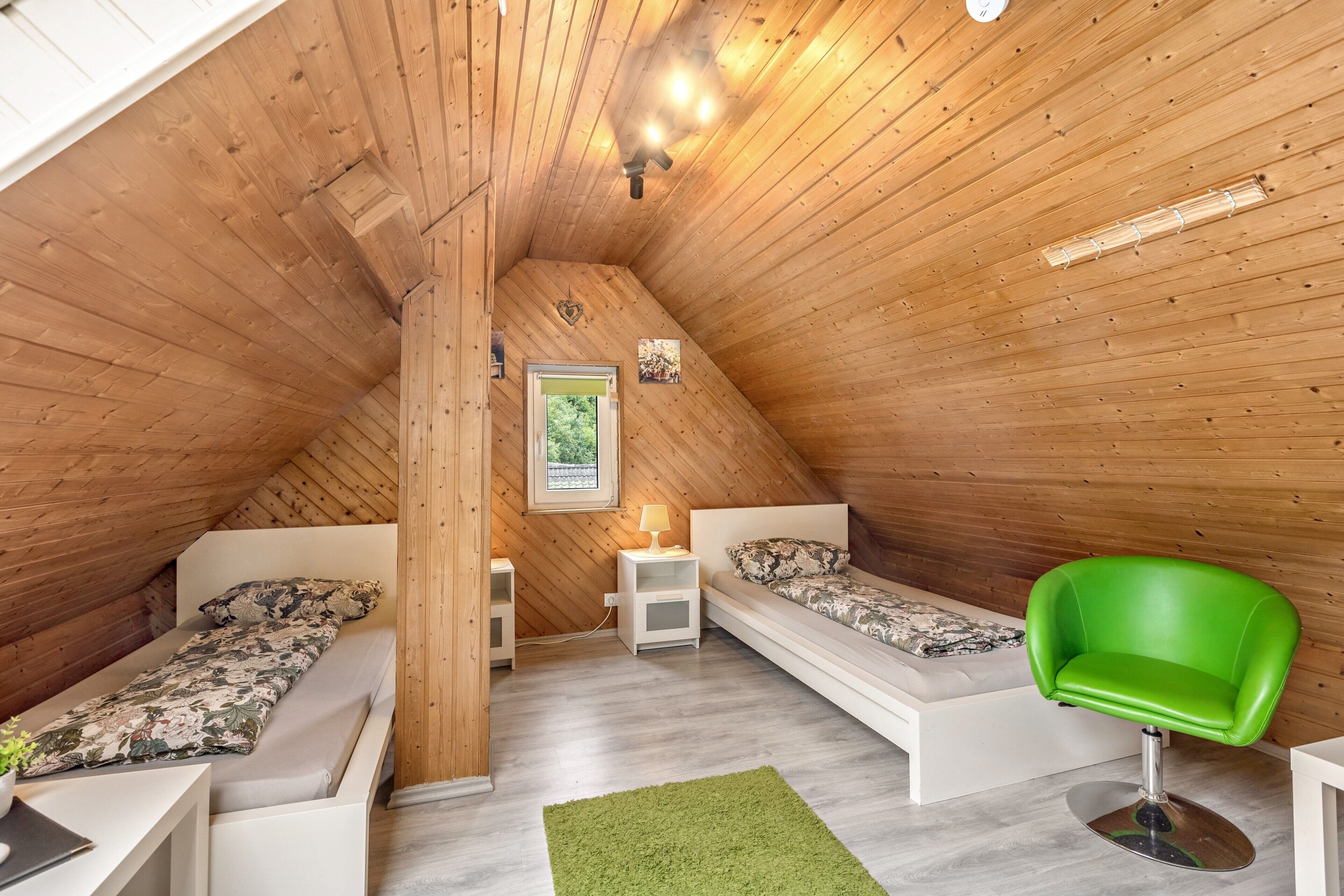 Une petite chambre mansardée en bois, avec deux lits, une chaise verte et une petite table.