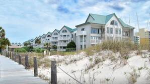 Un hôtel situé en bord de mer, composé de bâtiments à plusieurs étages, avec une promenade en bois menant à la plage et des dunes de sable.