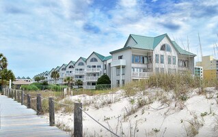 Un hotel frente a la playa con edificios de varios pisos, un paseo marítimo de madera que conduce a la arena y dunas de arena.