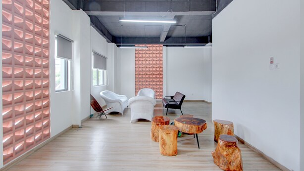 Un espace de bureau moderne avec un plancher en bois, des murs blancs et un mur orné d’un motif géométrique. On y trouve des chaises blanches et une chaise noire, ainsi que des tabourets en bois.