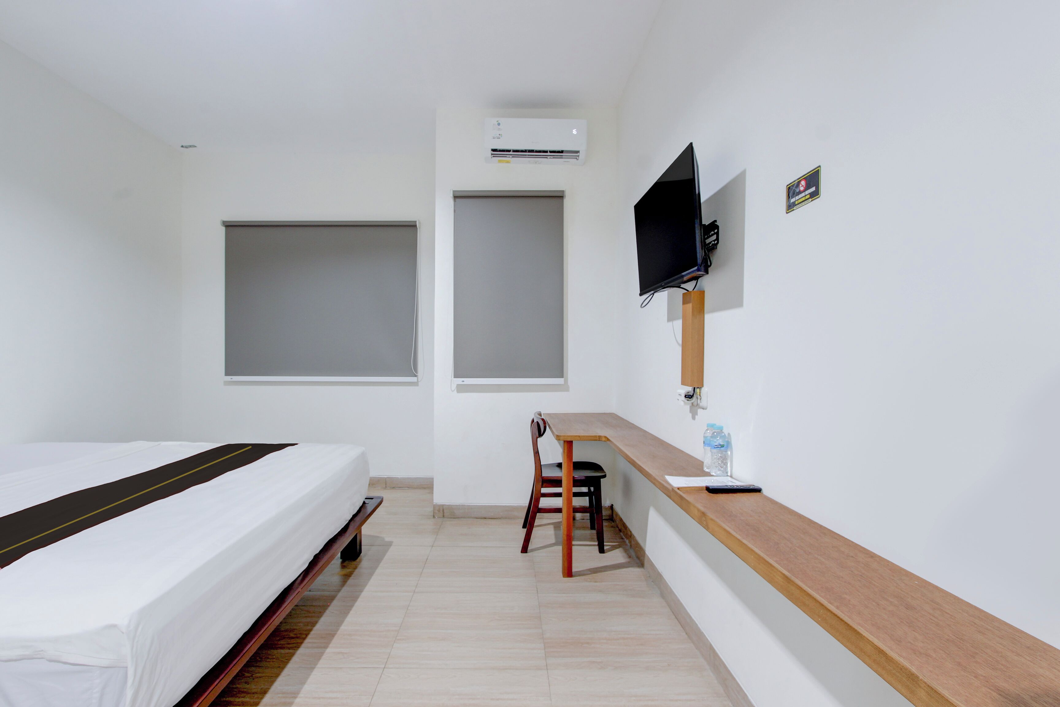 A hotel room with a bed, a desk with a chair, a TV mounted on the wall, and an air conditioning unit.
