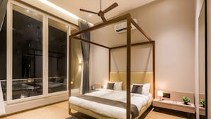 A modern bedroom with a four-poster bed, a bedside table with a lamp, and a view through large windows. - Suruchi Villa By Soumils Stays (Alibag)