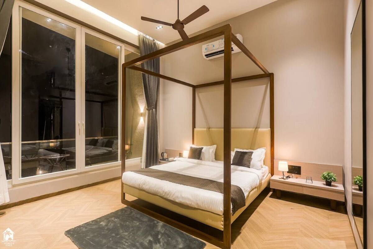 A modern bedroom with a four-poster bed, a bedside table with a lamp, and a view through large windows.