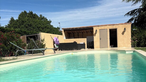 En swimmingpool med ett trädäck i förgrunden, en byggnad med en dörr och fönster i bakgrunden och träd som omger området.