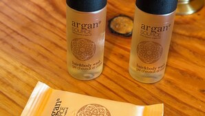 Två flaskor av Argan Source-schampo och duschgel för hår och kropp samt en påse med Argan Source-tvål, placerade på en träyta.