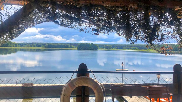 A view from a covered deck overlooking a calm lake, with a lifebuoy hanging on the railing and a bench in the foreground.