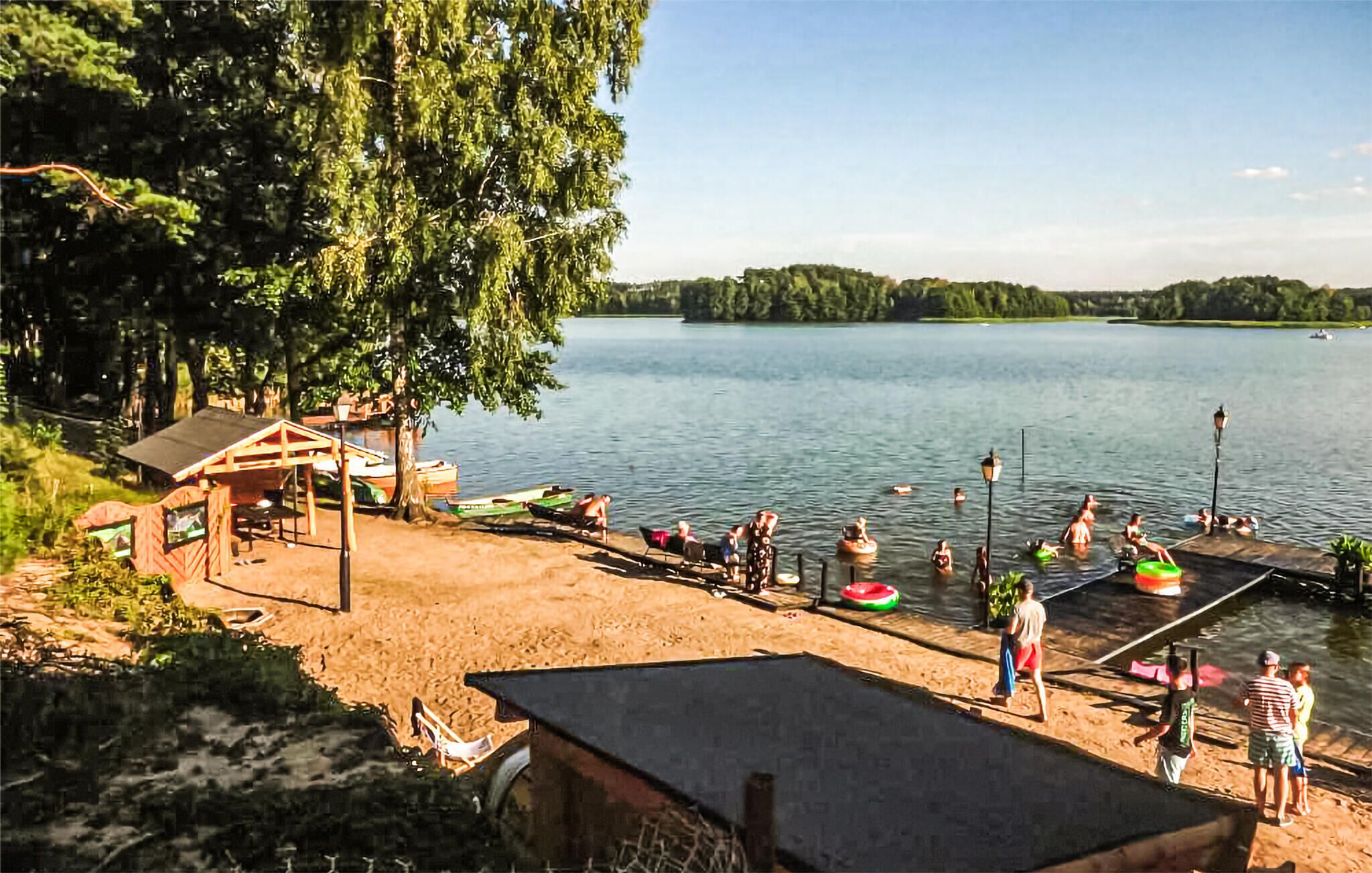 A lakeside beach with a wooden structure, boats, and people swimming.
