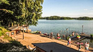 A lakeside beach with a wooden structure, boats, and people swimming.