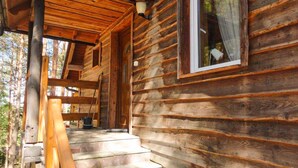 A log cabin with a porch, stone steps, and a window with a curtain.