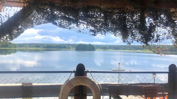 A view from a covered deck overlooking a calm lake with a distant island, a lifebuoy, and a small boat.