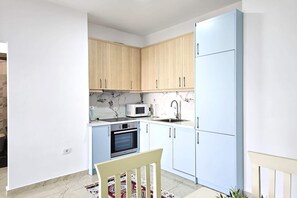 A compact kitchen with light wood cabinets, a white countertop, a microwave, an oven, and a sink. - One Bedroom Apartment In Saranda - 001 (Sarandë)