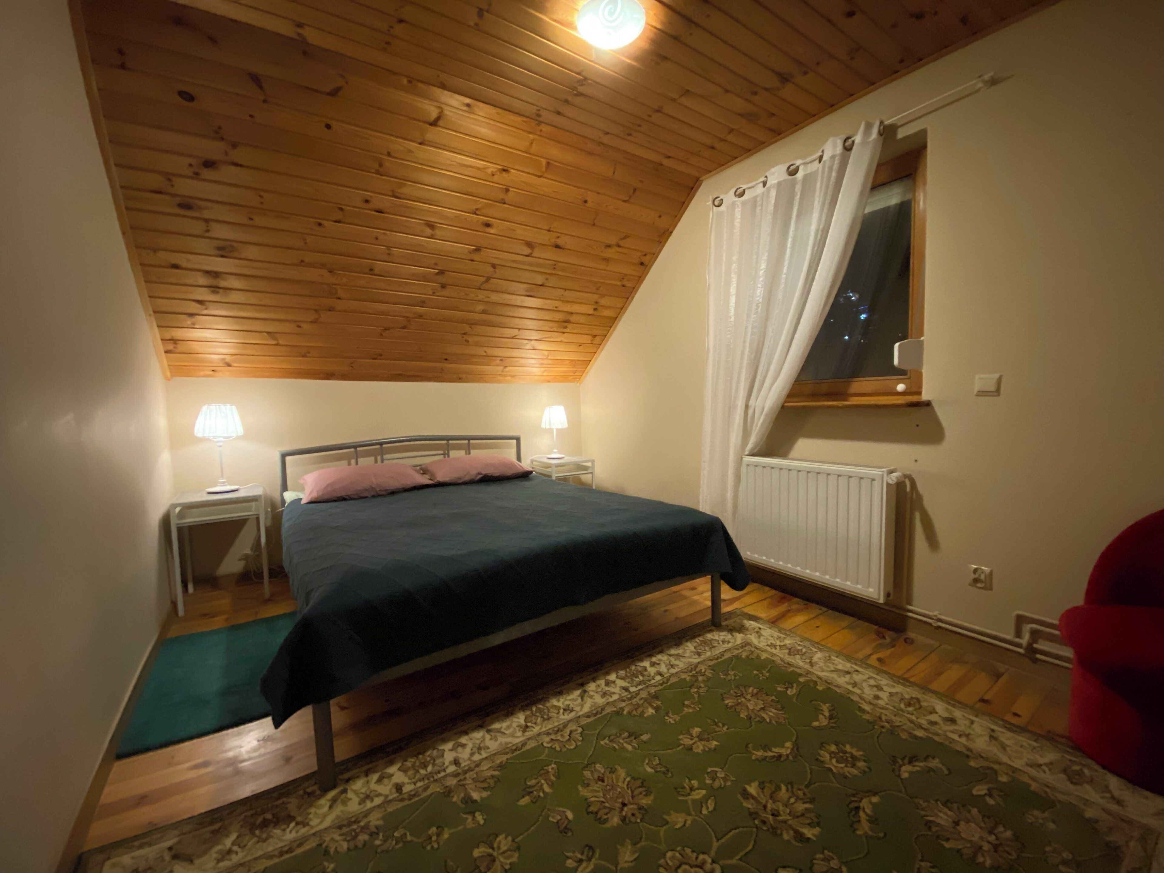 A bedroom with a wooden ceiling, a bed with a dark blue bedspread, two bedside lamps, a window with white curtains, and a green patterned rug.
