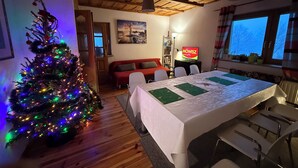 Ein gemütliches Zimmer mit Holzbalkendecke, einem Weihnachtsbaum mit bunten Lichtern, einem Esstisch mit Stühlen, einer Couch und einem Fernseher.